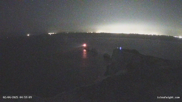 A dimly lit scene at dawn showing a coastline with faint lights in the distance. In the foreground, a blurred shape, possibly a boat, emits a red light reflecting on the water. The background reveals a horizon with muted lighting, suggesting a nearby town. The overall atmosphere is dark with a night sky, scattered stars, and minimal visibility. The timestamp at the bottom left indicates the date and time as February 4, 2025, at 4:59 AM.