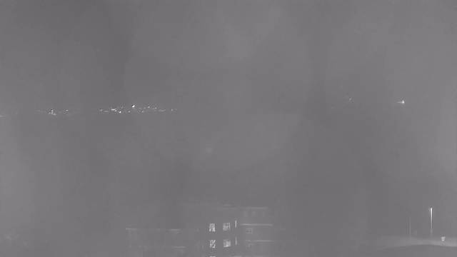 A dimly lit night scene showing faint lights in the distance, possibly from buildings or streetlights. In the foreground, there are indistinct shapes that may represent a structure, partially illuminated from a light source. The overall image has a grayish tone with a blurred appearance, suggesting low visibility or fog.