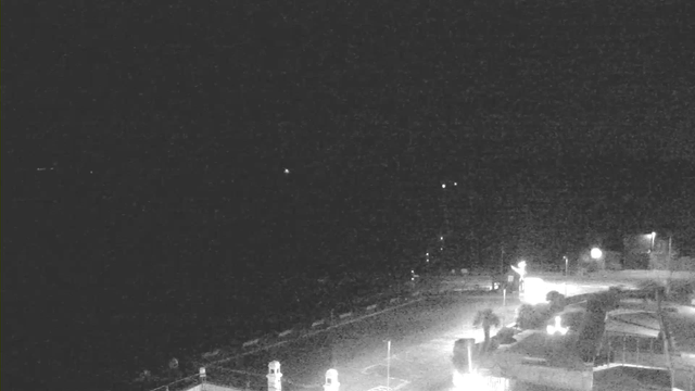 A dark scene captured by a webcam, showing very little visibility. The foreground features a walkway along a body of water, with faint outlines of structures, possibly buildings or a pier. Sparse lights can be seen in the distance, indicating the presence of nearby locations. The sky is predominantly black, and the overall atmosphere is dim and quiet.