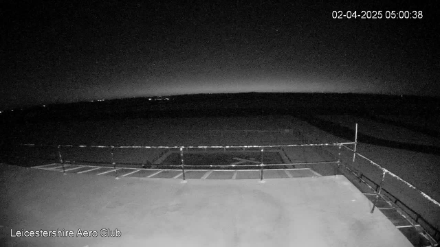 A dimly lit outdoor scene captured by a webcam, showing a flat surface with a railing on the left side. In the background, the horizon is barely visible against a dark sky, with faint lights scattered at a distance. The image appears to be taken early in the morning or late at night, indicating low visibility conditions. The date and time stamp indicates it is April 2, 2025, at 5:00 AM.