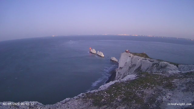 The image shows a coastal landscape at dawn. In the foreground, there is a rocky cliff with greenery. Below the cliff, the sea is calm and reflects a gradient of blue hues. In the distance, a series of white chalky rock formations emerge from the water. A red lighthouse is visible on one of the rocks, along with a small building nearby. The sky is clear with soft light, gradually brightening as the sun rises.