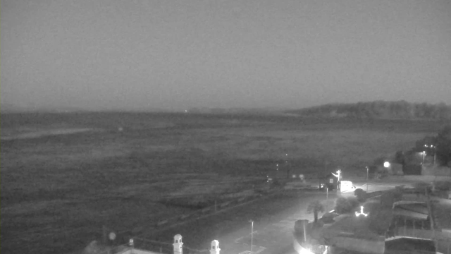 A dark, grainy webcam image depicting a vast, empty landscape with a body of water and a distant shoreline. The scene appears to be in low light, suggesting early morning or dusk. In the foreground, there are structures or buildings with soft illumination, and a winding road leads toward the water. The sky is uniformly dark, blending into the horizon where it meets the water. There are faint lights visible on the distant shore, adding subtle highlights to the evening scene.
