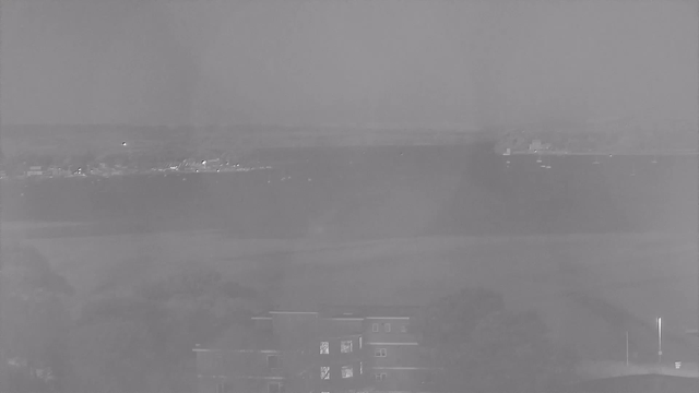 A grayscale image showing a view of a body of water in the distance, with some small boats visible on the water. In the foreground, there is a building with multiple windows. The landscape is mostly flat, with trees partially visible at the bottom of the image. The sky appears overcast or hazy, creating a muted atmosphere.