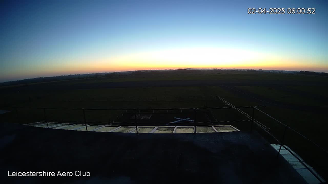 A scenic view from a webcam at the Leicestershire Aero Club, capturing the sky at dawn. The horizon displays a gradient of colors, transitioning from dark blue at the top to a soft orange and yellow near the horizon. Below, the foreground shows a flat, open field, with a light structure outlined in the lower left corner. There is a hint of a runway or landing area marked with white lines, while the overall atmosphere conveys a serene, early morning setting.