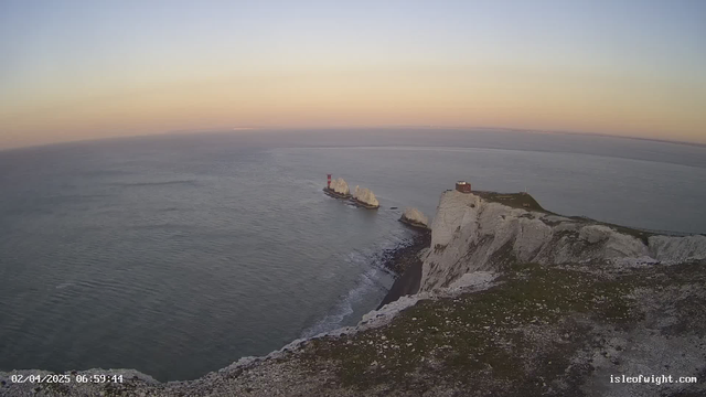A scenic view of a coastline at sunrise, featuring white cliffs on the right with patches of grass and rocks. To the left, a red lighthouse stands on a rocky outcrop extending into the calm sea, which reflects shades of blue and pink from the sky. The horizon is faintly visible in the distance, where the sky transitions from light to darker hues. A small building is situated at the edge of the cliffs.