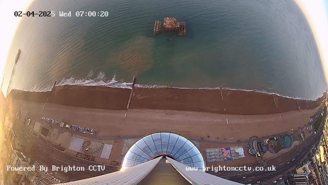 Aerial view from a high structure overlooking a beach at dawn. The sandy beach stretches along the bottom with a row of wooden structures, likely piers, extending into the calm sea. Waves gently lap at the shore. The water has a gradient of blue to green hues. In the foreground, a circular glass roof structure is visible, with buildings and amusement rides along the coastline. The scene is softened by early morning light, and a timestamp in the corner indicates the date and time.