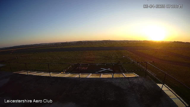 A panoramic view of a grassy airfield at sunrise. The sky transitions from deep blue at the top to warm orange and yellow near the horizon. Below, an empty airstrip and a small white airplane shape are visible on the ground. In the foreground, there is a railing along a flat surface, suggesting a vantage point from a building. The date and time are displayed in the corner.