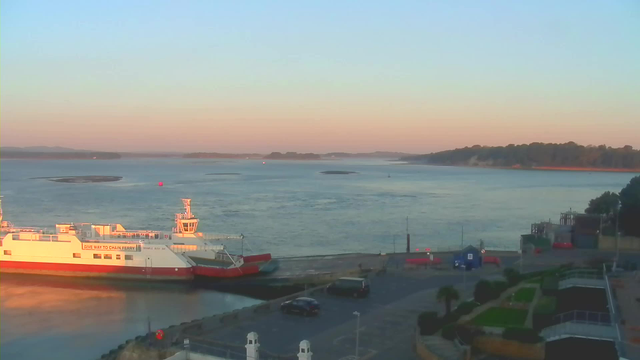 A calm body of water reflects soft colors of dawn with light blue skies above. To the left, a white and red ferry is docked near a concrete pier, with the words "GIVE WAY TO CHAIN FERRY" visible on its side. In the foreground, there are parked vehicles and a few palm trees. The right side features a small blue building and additional structures, while distant hills are visible in the background.
