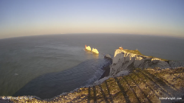A coastline view is captured, featuring white chalk cliffs rising over the blue sea. In the foreground, rocky terrain and patches of grass are visible. Off the coast, two prominent white rock formations are apparent, with a red beacon standing on one of them. The sky is clear, transitioning from light blue to pale hues, and the sun is low in the sky, casting long shadows on the cliffs. The image evokes a serene coastal landscape.