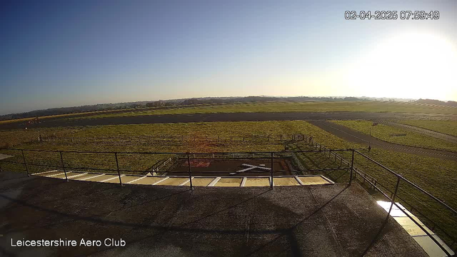 A view from a webcam at Leicestershire Aero Club showing a clear sky with the sun rising in the upper right corner. Below, there is a large grassy area leading to an empty runway. The grass is green and well-maintained, with a fence outlining a section near a helipad marked by a white cross. In the background, rolling hills are visible, and a tarmac path runs through the scene. The image captures a tranquil morning atmosphere, highlighting the beauty of the landscape.