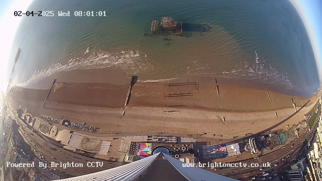 A panoramic view of a beach during early morning. The sandy shoreline is visible, with gentle waves lapping at the edge. In the background, there is an old pier structure partially submerged in the water. To the right, the beachfront features several buildings, including amusement attractions and colorful umbrellas. The image is taken from an elevated position, looking downwards toward the shoreline. The time and date are displayed in the top left corner.