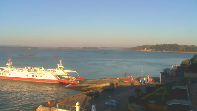 A large white and red ferry is docked at a concrete pier by the water. The calm sea reflects the clear sky, which is a pastel blue near the horizon. In the background, there are some small islands and a shoreline with trees. On the pier, there are a few parked cars and some structures, including a building. Bright markers are visible on the water, indicating navigational points. The scene captures a peaceful coastal setting in the early morning light.