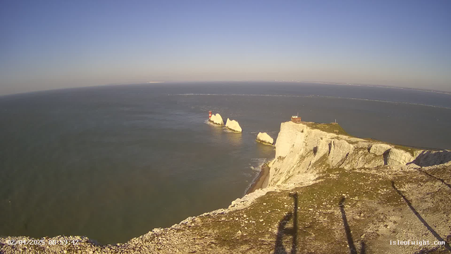 A coastal view shows white chalk cliffs on the right side of the image, leading towards the ocean. In the water, two small islands are visible; one with a red lighthouse and the other with a structure. A clear blue sky occupies the upper part of the image, with the horizon blending into the sea. The foreground features rocky terrain.
