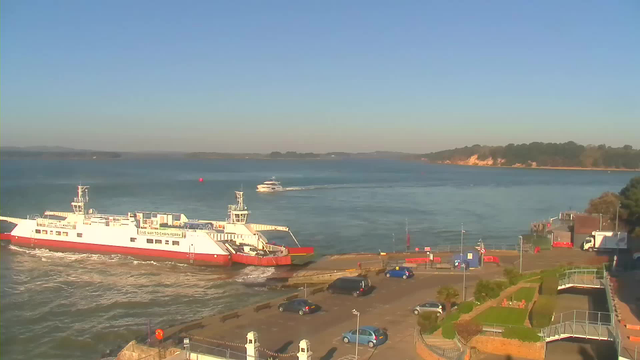 A scenic view of a waterfront with a white and red ferry docked at the pier. The ferry is surrounded by water, with ripples visible. In the background, a small motorboat is navigating across the water, while a green hill with trees and a sandy shore are discernible in the distance. The foreground shows a parking area with several cars and landscaped greenery, including palm trees and pathways leading down to the water. The sky is clear and blue, indicating a sunny day.