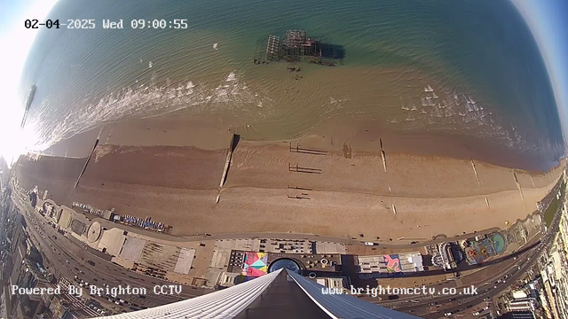 An aerial view of a beach with golden sand and gentle waves lapping at the shore. In the water, there is a partially submerged pier structure. The beach is mostly empty, with a few scattered people. There are colorful beach umbrellas and a vibrant amusement area visible near the shore. The image shows a clear blue sky and the time and date displayed in the upper left corner.