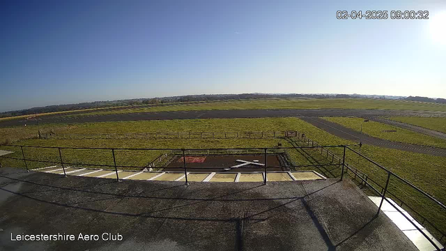 A clear blue sky is visible with no clouds. In the foreground, a railing lines a flat roof area with a small helipad marked by a white cross on a red background. Below, a grassy field stretches out, bordered by a wooden fence. A paved runway is visible further back, with additional grassy areas and a few distant hills on the horizon. The scene is bright and well-lit, indicating daytime. The timestamp displayed shows 9:00 AM on April 2, 2025.