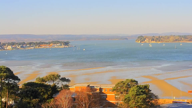 A panoramic view of a calm bay with a sandy beach. In the foreground, there are a few buildings partially obscured by trees, with a variety of green foliage. The water is a mix of blue and turquoise, reflecting the clear sky above. Several small boats are scattered across the water, while a distant shoreline reveals a cluster of houses and a hill with trees. The background features rolling hills that fade into the horizon.
