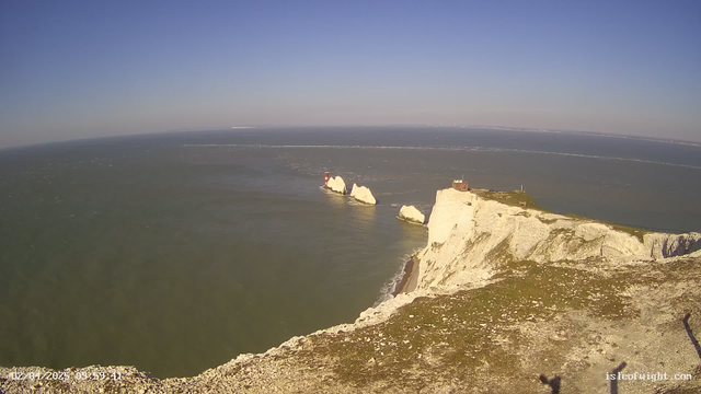 A panoramic view of a coastal landscape featuring white chalk cliffs on the right side, overlooking a blue sea. Two lighthouses are positioned on rocky outcrops in the water. The sky is clear and sunny, with a few wispy clouds. The ocean has gentle waves and visible ripples, indicating a light breeze. The foreground shows grassy terrain with small stones and a rugged cliff edge.