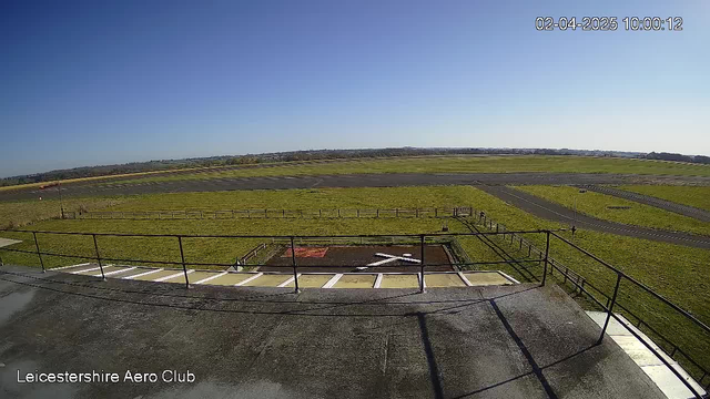 A clear blue sky is visible overhead, with a large expanse of green grass and a runway extending into the distance. In the foreground, there is a flat rooftop with a railing, and a marked helipad with an X symbol on it. The area is surrounded by a fence, and a small road can be seen leading off the edge of the runway. The scene appears to be at an airfield associated with the Leicestershire Aero Club.