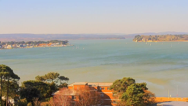 A scenic view of a body of water with gentle waves, surrounded by trees and land in the background. In the distance, a small town is visible along the shoreline with various buildings and boats, while hills rise in the far background. The sky is clear with a light blue hue, indicating a sunny day. The foreground features a cluster of trees and a building that has a reddish-brown exterior.