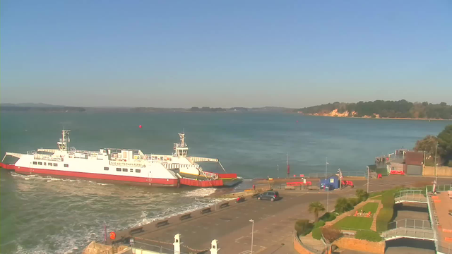A white and red ferry is docked at a harbor with gentle waves surrounding it. The background features calm blue water and a clear sky, while a small coastline with green trees and sandy cliffs is visible in the distance. In the foreground, there is a parking area with a few cars and a landscaped area with plants.