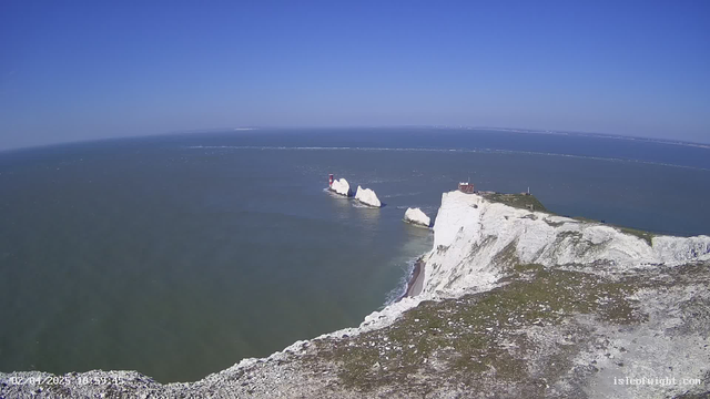 A panoramic view of a coastline featuring white chalk cliffs alongside a blue sea under a clear sky. In the distance, there are two small lighthouses on rocky outcrops. The foreground shows a grassy area atop the cliff, leading down to the water. Light waves create ripples on the surface of the sea, and a faint horizon line is visible where the sea meets the sky.