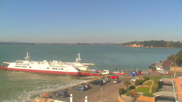 A ferry is docked at a terminal on the edge of a calm body of water, under a clear blue sky. In the foreground, there are several parked cars and a small road leading up to the terminal. The shoreline in the background features trees and a sandy beach, with a slight elevation. A few boats can be seen on the water in the distance. The scene is bright and sunny, creating a peaceful, maritime atmosphere.