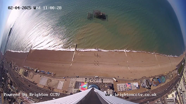 A high-angle view of a beach scene on a sunny day, showing the ocean meeting the shoreline. The water is a blue-green color with gentle waves lapping at the sandy beach. People can be seen walking along the shore, with some standing by the water's edge. There are several piers extending into the water, and structures along the beach, including colorful canopies and attractions. In the lower part of the image, there is a waterfront promenade with a variety of shapes and colors visible. The sky is clear with bright sunlight reflecting off the water.