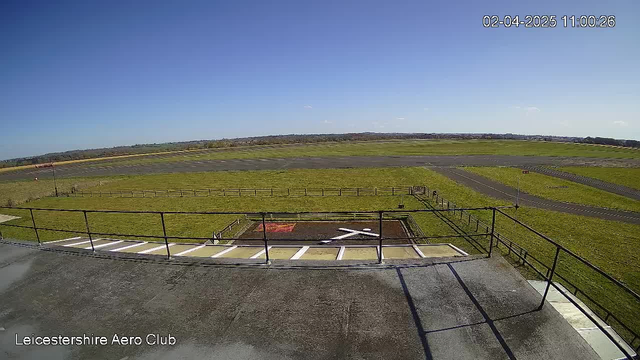 A clear blue sky stretches across the top of the image. Below, a wide open space of greenery and a runway can be seen. In the foreground, there is a flat rooftop with a railing. A helipad marked with a large white "X" is visible on the ground, surrounded by grass and a few fences. The scene conveys a bright, sunny day at the Leicestershire Aero Club, with no clouds in sight.