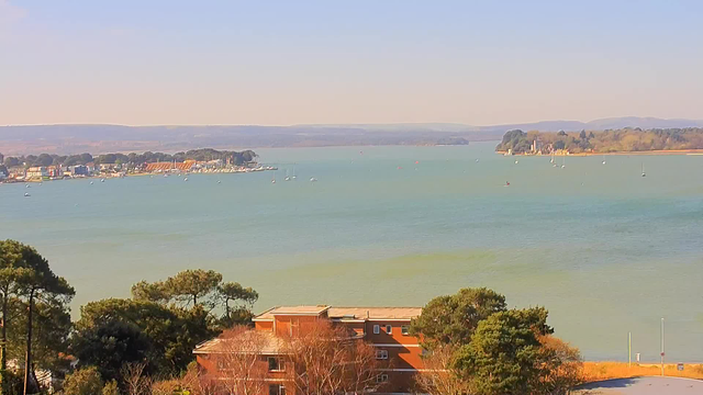 A scenic view of a calm waterway with several small boats scattered across the surface. In the foreground, there are some trees and a low building with a reddish-brown facade. Along the shoreline, colorful buildings are visible, leading to a gentle slope of land that extends toward the horizon. In the background, faint hills and trees outline the farthest edge of the scene under a clear blue sky, creating a peaceful coastal atmosphere.