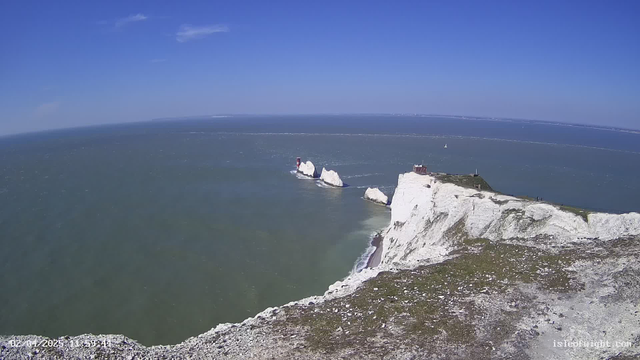A scenic view of a coastline featuring prominent white cliffs and a blue ocean. In the distance, several narrow, rocky formations rise from the water, and two red lighthouses are positioned on these rocks. The sky is clear with a few white clouds, and there is a small sailboat visible on the water. The foreground includes a patch of grassy land at the cliff's edge.
