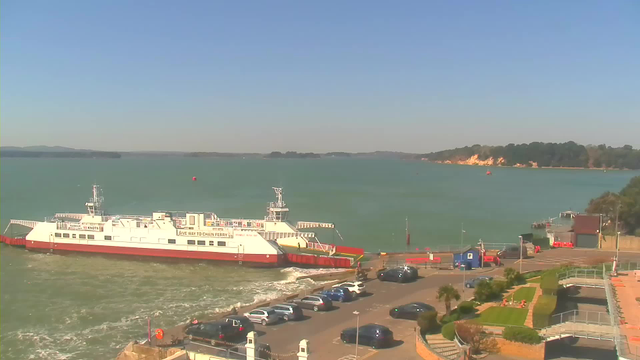 A ferryboat is docked at a waterfront terminal with a clear blue sky overhead. The ferry, painted white and red with signage indicating it operates at "10 knots," is positioned near the shore. In front of the ferry, there is a parking lot filled with various cars. To the right, lush greenery and landscaped areas are visible, along with a small building and additional waterfront structures. The calm water extends into the distance, where low hills and trees line the horizon.