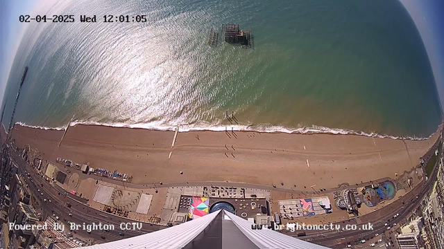Aerial view of a sandy beach with gentle waves lapping at the shore. There are a few small figures walking along the beach. A pier is visible in the distance, partially submerged in water. Below, colorful displays and structures can be seen along the promenade. The ocean is a gradient of greens and blues, reflecting bright sunlight. The image has a timestamp in the top left corner indicating the date and time.