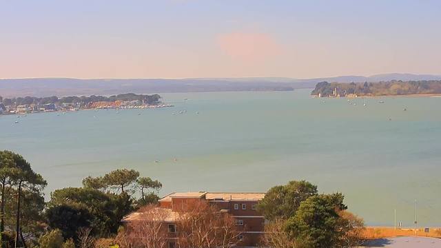 A panoramic view of a calm bay with turquoise water. In the foreground, there are patches of green trees and a low-rise building with brick exterior. The midground features several small boats scattered across the water, while a beachfront with houses and sailboats lines the shore on the left side. In the background, low hills rise gently towards the horizon under a clear, light blue sky.