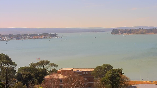 A serene view of a calm bay, with clear turquoise water and scattered small boats. In the foreground, there are trees and a low building. The shoreline features a mix of houses and a marina with more boats visible. In the distance, green hills rise gently along the horizon under a light blue sky.