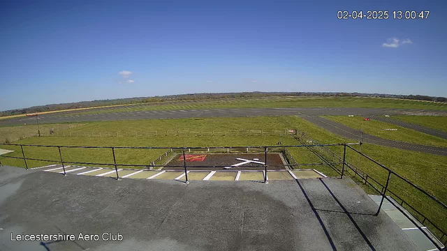 A view from a camera positioned on a structure overlooking an airfield. The foreground shows a flat rooftop with a railing. Below, there is a grass field and runway, which is divided into sections. A helipad is visible in the lower left corner, marked with a red square and white cross. In the background, there are distant green hills beneath a clear blue sky with a few scattered clouds. The image is bright and captures a sunny day.