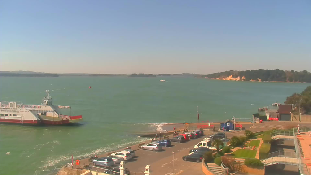 A scenic view of a waterfront area under a clear blue sky. In the foreground, there is a parking lot with multiple parked cars of various colors. Near the water, a ferry is docked, with its bow facing towards the viewer. Just beyond the ferry, smaller boats are visible navigating the water, which appears a vibrant teal. The shoreline features trees and a sandy area with some structures. In the distance, there are hills and a sandy cliff. The scene is bathed in bright sunlight, creating a warm ambiance.