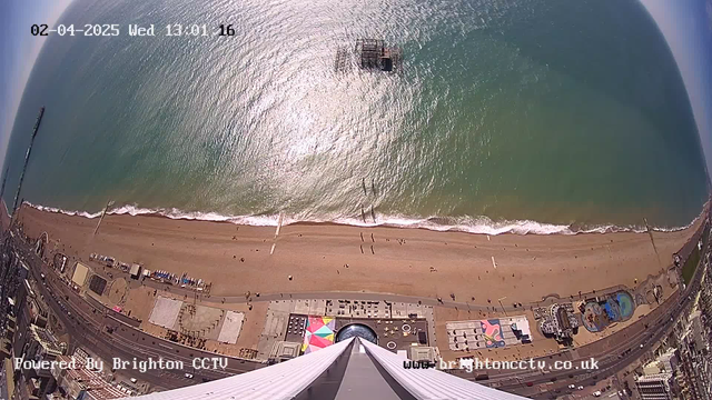 A high-angle view of a beach and ocean taken from a webcam. The sandy beach stretches across the lower portion of the image, with a few groups of people walking along it. In the ocean, gentle waves can be seen reflecting sunlight. Several beach structures, including a pier and amusement rides, are visible along the shore. The sky appears clear and blue, indicating a sunny day. The image includes a timestamp at the top showing the date and time, along with a watermark at the bottom identifying the camera source.