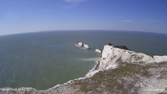 A coastal view showing white chalk cliffs rising above the sea. In the distance, several rocky outcrops are visible, with a red lighthouse on the left side. The ocean is a mix of green and blue hues, and the sky is clear with a few wispy clouds. The foreground features a grassy area on top of the cliffs, leading down to a beach near the water. The image captures a vibrant, sunny day.