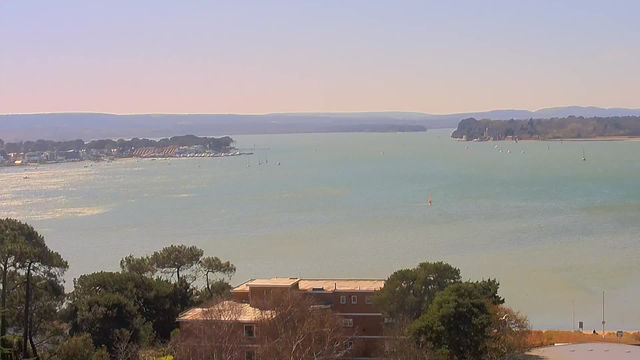 A panoramic view of a calm body of water under a clear sky, with distant hills visible on the horizon. Along the shoreline, several small boats are sailing, and a few houses and trees are seen in the foreground. The water reflects light, creating a shimmering effect.