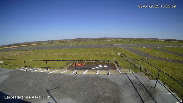 A clear blue sky is visible above a wide open field and a runway of an airfield. In the foreground, there is a railing and a helipad marked with a white cross and red paint underneath it. The ground is predominantly green grass, and the runway extends in the distance, indicating the layout of the airfield. The time and date are displayed in the corner of the image.