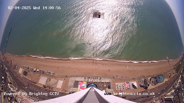 A high-angle view from a tower looking down at a beach and shoreline. The sandy beach stretches horizontally across the bottom of the image, with people walking and sunbathing. The ocean appears calm and sparkles in the sunlight. There are pier structures visible extending into the water. Below, various colorful structures are seen near the beach, including a large colorful tent, and a few buildings. The sky is clear with some light clouds. The date and time are displayed in the top left corner.