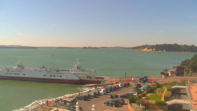 A ferry is docked at a harbor by the water, with gentle waves lapping against it. Several parked cars are arranged in a lot next to the ferry terminal. In the background, the sea stretches towards a horizon with distant, low hills and a shoreline lined with greenery and cliffs. The sky is clear and bright, with a few clouds visible.