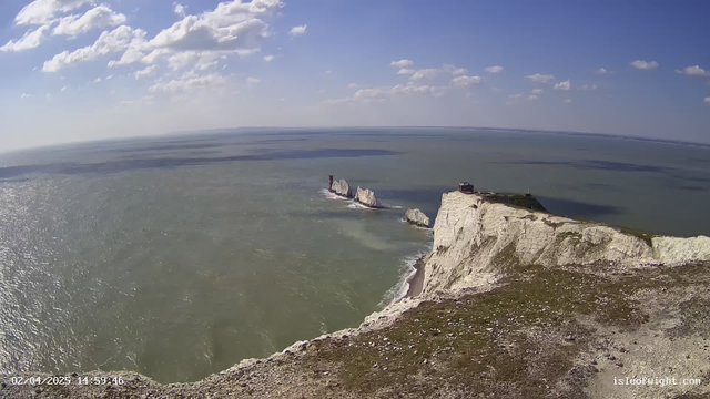 A wide view of a coastal landscape featuring white chalk cliffs, with a rocky formation emerging from the water towards the center of the image. The ocean is calm with rippling waves reflecting sunlight, and a blue sky filled with scattered white clouds stretches overhead. In the distance, the coastline continues, and a small structure is visible at the top of the cliffs. The foreground includes grassy terrain with scattered rocks and flora.