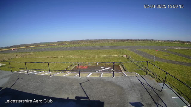 A clear blue sky fills the upper part of the image. Below, an expansive green landscape is visible, with a paved runway running horizontally across the center. To the left, a wind sock is seen indicating wind direction, while a grass area with a white 'X' marking is positioned in the foreground. A wooden fence borders the grassy area, and the scene appears to be peaceful and uncluttered, likely from a height showing a view of the Leicestershire Aero Club.