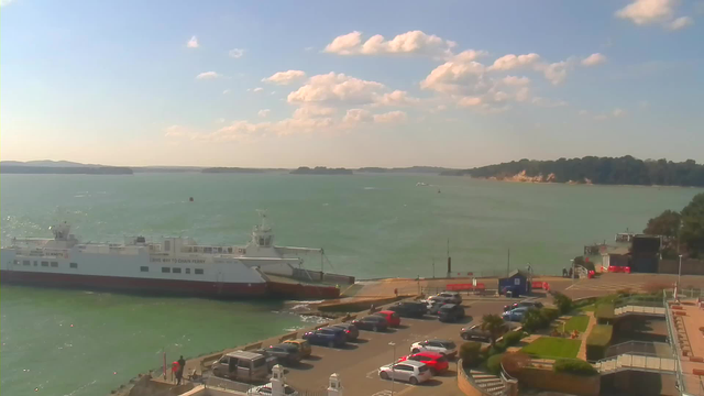 A sunny waterfront scene featuring a large white ferry docked at a terminal. In the foreground, there is a parking lot filled with various cars in different colors. The water is a greenish-blue, with gentle waves and a few boats in the distance. Some green trees and cliffs can be seen along the shoreline. Fluffy white clouds are scattered in a bright blue sky.
