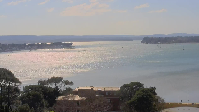 A serene landscape view featuring a calm body of water reflecting sunlight, creating a sparkling effect. In the foreground, there are some green trees and a building with a flat roof. The background shows a shoreline with scattered boats on the water, alongside hills in the distance under a clear sky with a few clouds.
