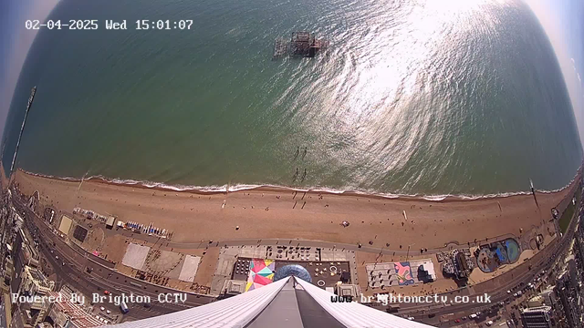 A wide-angle view of a sandy beach along a coastline, with the ocean gently lapping at the shore. People can be seen walking along the beach and some are sunbathing. In the distance, there is a pier extending into the water and a structure, possibly a raised platform or amusement area, situated on the beach. Colorful beach chairs are arranged on the sand, and there are various buildings lined along the street near the beach, including a circular structure and a large, colorful patterned area in front of one of the buildings. The time displayed in the corner indicates it is 15:01 on April 2, 2025. The sunlight reflects off the surface of the water, creating a shimmering effect.
