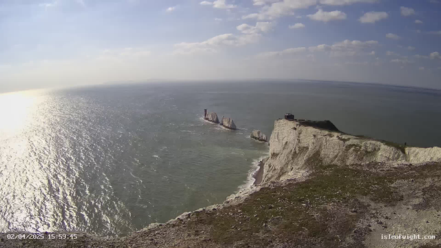 A panoramic view of a coastal landscape featuring a rocky cliff edge on the right side. The cliff is mostly white with patches of green grass. The sea extends across the image, reflecting sunlight and creating a shimmering effect. In the distance, several large rock formations rise from the water, with one notably tall and narrow structure. The sky is partly cloudy with blue and gray tones. A small building is visible on top of the cliff, offering a vantage point of the sea and the rocks below.