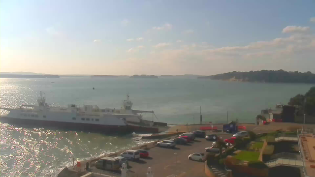 A view of a waterfront scene featuring a large ferry docked at a harbor. The water is calm and reflects sunlight, creating a shimmering effect. In the foreground, there is a parking area with several cars, and a small garden with green plants. The background shows distant islands on the horizon under a clear sky with a few scattered clouds.